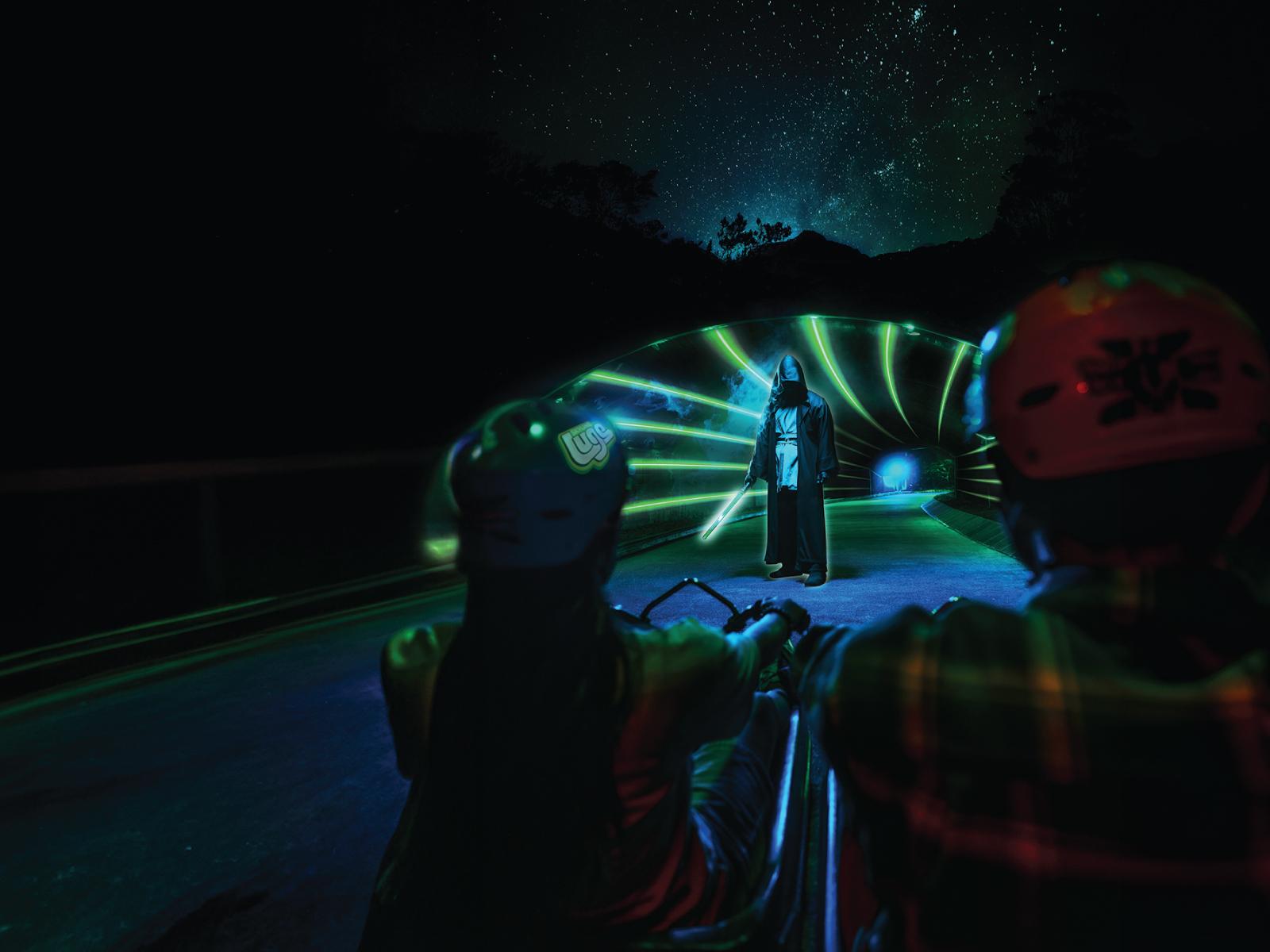 Two people Luge through an immersive light tunnel seeing a person holding a lightsabre.